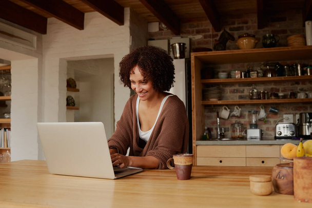 Confident biracial young woman in casual clothing working from home on laptop in kitchen - Photo, image