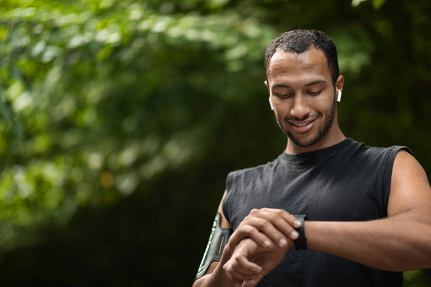 Closeup of handsome young black sportsman exercising in public park, setting fitness tracker before workout, checking heart rate, panorama with copy space. Modern technologies and sport concept - 写真・画像