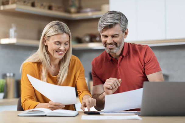 Loving spouses attractive middle eastern man and woman sitting at kitchen table in front of computer, holding papers, using calculatir, working on family budget together, copy space - Photo, image