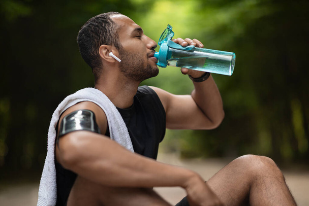 Athletic young black man drinking water while having break, training at public park, using wireless earbuds, listening to music while exercising, copy space. Hydration and sport concept - 写真・画像
