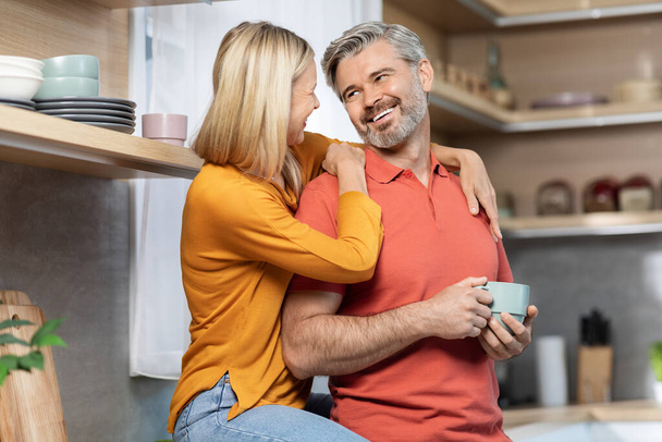 Loving blonde lady sitting on table, hugging her smiling middle aged husband, drinking morning coffee together and having conversation at kitchen at home, enjoying time together, copy space - Фото, изображение
