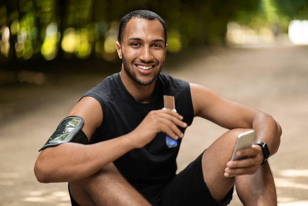 Hungry african american sportsman eating protein bar while resting, having workout at public park, sitting on the ground, using cell phone and smiling, watching fitness video, copy space - Fotoğraf, Görsel