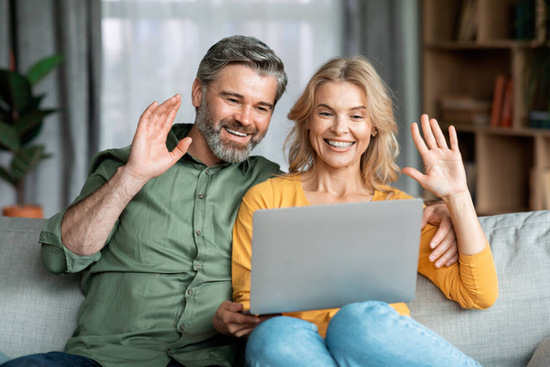Happy Middle Aged Couple Making Video Call With Laptop At Home, Cheerful Mature Spouses Waving Hands At Webcam While Sitting On Couch In Living Room, Enjoying Online Communication, Closeup - Zdjęcie, obraz