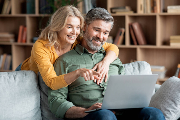 Happy Middle Aged Couple With Laptop Ordering Things From Internet Together While Relaxing On Couch In Living Room, Smiling Mature Spouses Making Virtual Shopping Or Booking Hotel Online, Closeup - 写真・画像