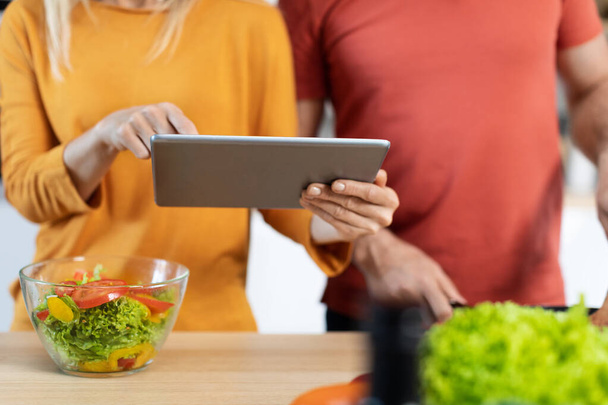 Cropped of man and woman using digital pad while cooking, unrecognizable couple preparing delicious healthy meal, reading food blog on Internet or watching video, closeup photo - 写真・画像