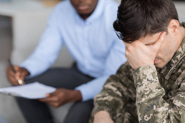 Вид зверху на молодого чоловіка у військовій формі солдата, який сидить на дивані, торкається голови під час бесіди з чорним лікарем, ветеран, який відвідує психотерапевта в клініці
 - Фото, зображення