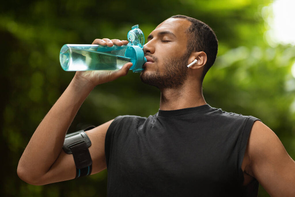 Athletic young black man drinking water, training at public park, using wireless earbuds, listening to music while jogging by forest, closeup shot. Hydration and sport concept - Foto, imagen