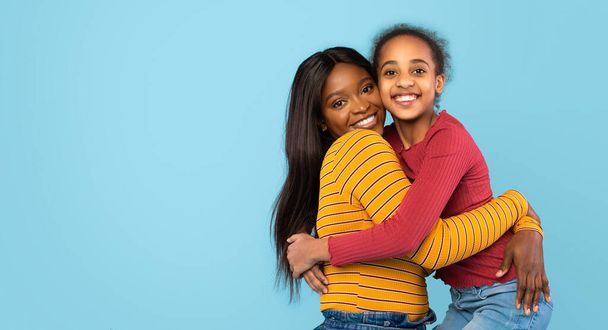 Love and care. Pretty african american daughter hugging her young happy mother, standing over blue studio wall, smiling at camera, expressing tenderness, panorama with free space - Foto, Imagem