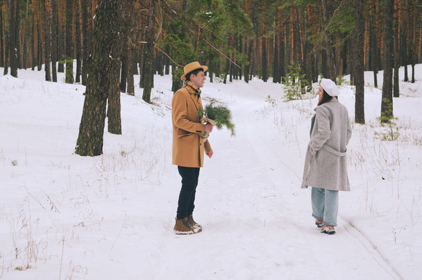 Portrait of funny family, couple in snowy park, forest. Young woman, girl and man in hat walking near snow covered pine trees. Drinking coffee. Having fun. Family winter holidays and date. Love - 写真・画像