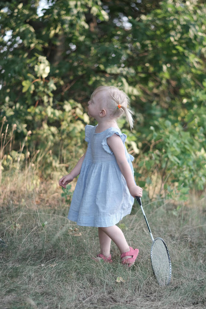 Blonde little girl with two tails and summerdress plays badminton. Child holds racket and shuttlecock. Summer vacation. - Foto, Imagen