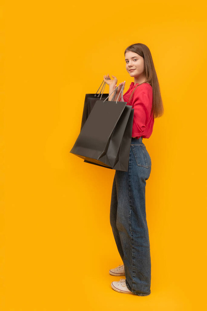 Beautiful young girl holds black paper bags after shopping on yellow background. Black Friday Concept. Vertical frame - Valokuva, kuva