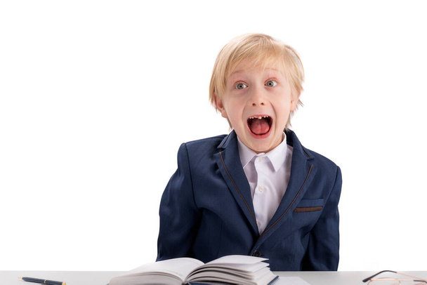 Schoolboy fools around while doing homework. Blond boy in school uniform makes grimaces. Screaming fidget boy. Isolated on white background - 写真・画像
