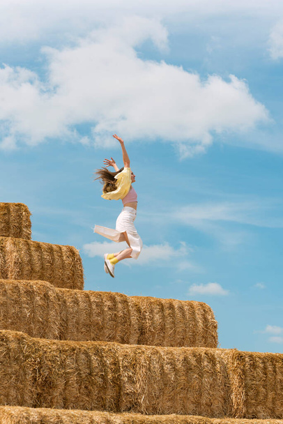Girl jumps on the haystack with her hands raised high. Countryside holiday on the farm. Hay harvesting - Fotoğraf, Görsel
