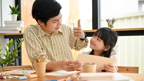 Handsome and kind Asian male art teacher giving a thumbs up to his adorable young student. Asian dad and daughter enjoys painting together. - Foto, Bild