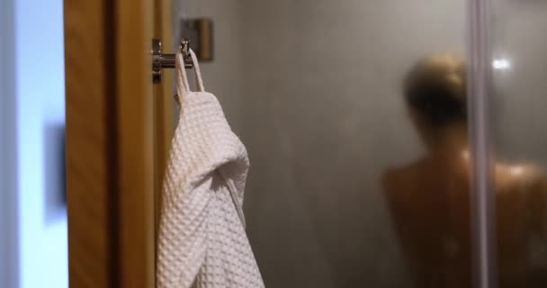 Naked woman is washing in shower and white bathrobe on hook. Evening and morning body hygiene - Imágenes, Vídeo