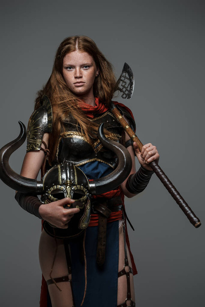 Shot of female warrior with helmet and axe dressed in armor against grey background. - Foto, immagini