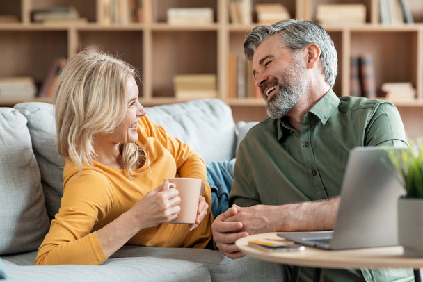 Portrait Of Happy Middle Aged Spouses Chatting And Drinking Coffee At Home, Romantic Mature Couple Resting On Couch, Looking At Each Other And Smiling, Enjoying Spending Time Together, Closeup Shot - 写真・画像