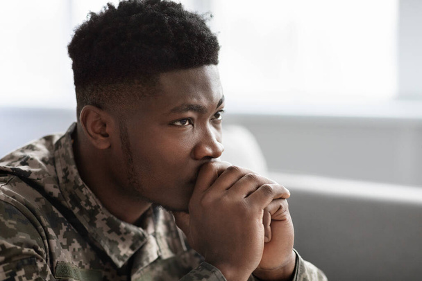 Side view of thoughtful military black man staring at copy space, holding palms by mouth, sitting on couch at home. Soldier visiting psychologist, suffering from posttraumatic stress, closeup - Фото, изображение