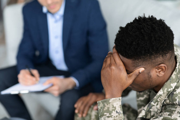 Top view of pensive african american man in military uniform soldier sitting on couch, touching his head while having conversation with doctor, veteran attending psychotherapist at clinic - Fotó, kép