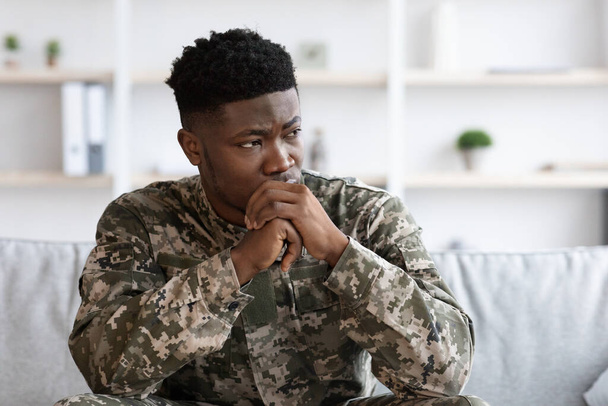 Depressed military black man staring at copy space, holding palms by mouth, sitting on couch at psychotherapist office. Soldier visiting psychologist, suffering from posttraumatic stress, closeup - Фото, зображення