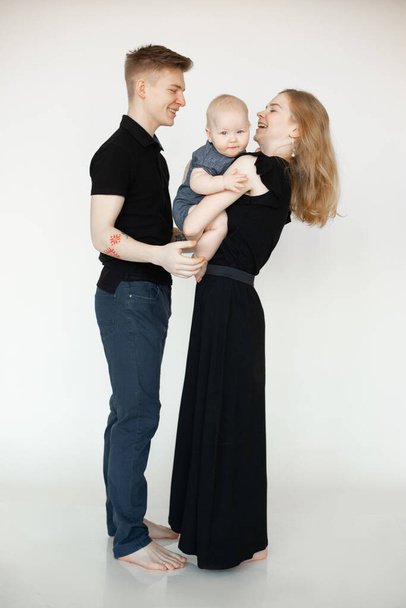 Portrait of young smiling beautiful family in dark clothes standing with little plump grey-eyed baby infant on white background. Mother holding child with hands. Family, relationship, having fun. - Fotó, kép