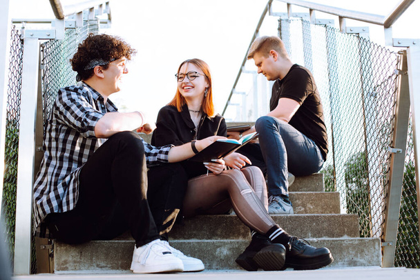 Cheerful talking relaxing and joyful small group of people, two men and red hair woman in glasses sitting on stairs and reading book. Three young friends on meeting weekend, vacation. Hipster culture - 写真・画像