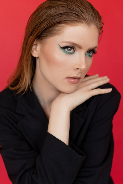 Vertical close up of young blonde woman in black capote shirt looking away, leaning on hand and posing like fashion model on red studio background. Professional make up. Mascara, eyeliner and shades - Φωτογραφία, εικόνα