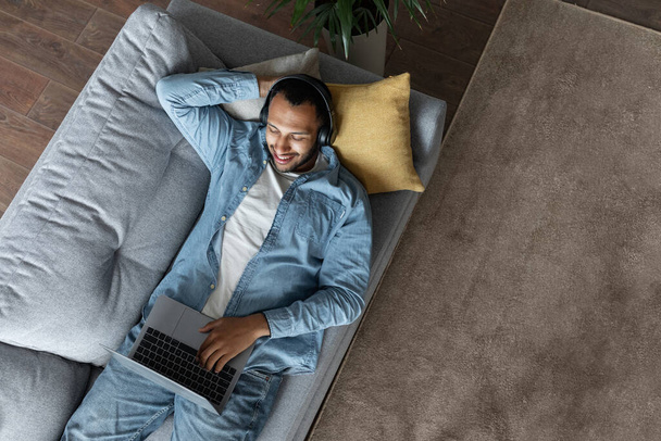 Top View Shot Ot Young Black Man Wearing Wireless Headphones Relaxing On Couch With Laptop, Handsome African American Guy Resting On Sofa At Home With Computer, Browsing Internet, Copy Space - Foto, Bild