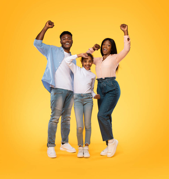Victory concept. Excited african american family of three shaking clenched fists, making winner gesture over yellow background. Happy black man, woman and girl celebrating success together - Fotografie, Obrázek