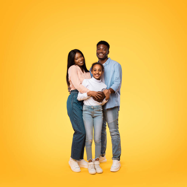Portrait of happy black family of three embracing and posing together over yellow studio background. Young african american parents hugging their daughter and smiling at camera - Zdjęcie, obraz
