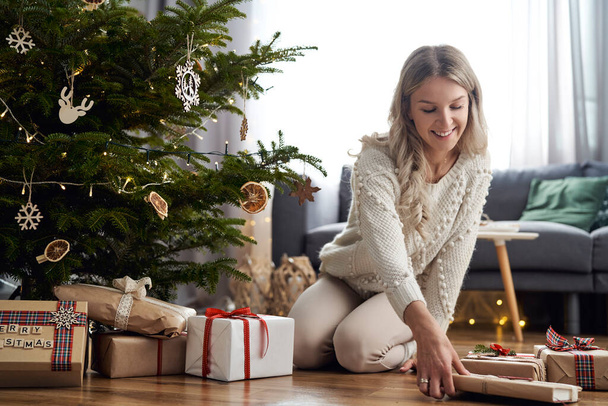 Young caucasian woman putting Christmas gifts under Christmas tree - Foto, Imagen
