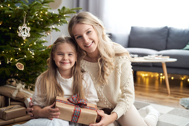 Portrait of caucasian girl and mother embracing each other and holding Christmas gift - Foto, immagini
