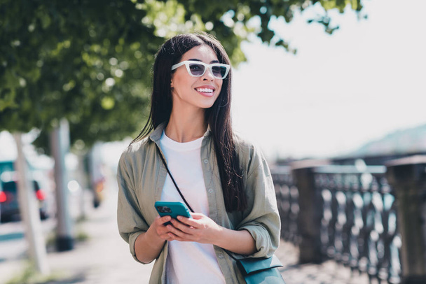 Photo of sweet dreamy lady dressed casual clothes dark glasses walking chatting modern gadget sunshine outside urban embarkment. - Foto, Imagem