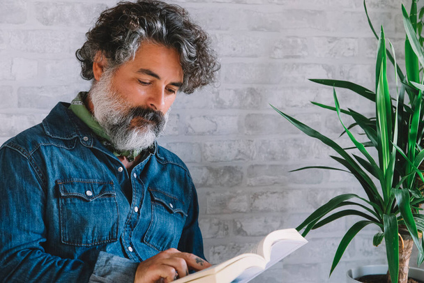 A handsome, bearded man wearing a denim shirt. Man sitting on a chair, reading a book and relaxing. - Foto, Imagen