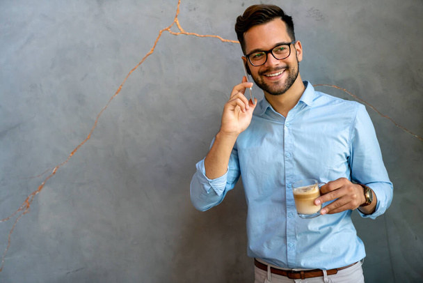 Portrait of a smiling young handsome business man using smartphone and holding cup of coffee - Φωτογραφία, εικόνα