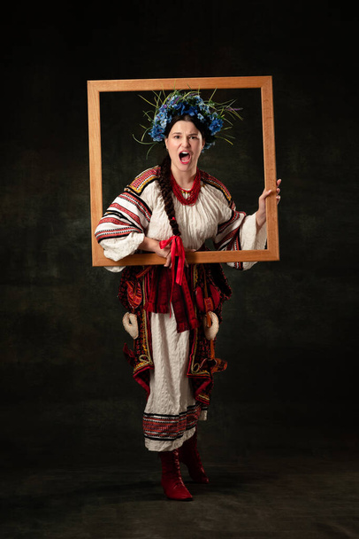Independence. Emotional young woman wearing national folk Ukrainian attire isolated over dark retro background. Human emotions, history, ad, fashion, beauty, eras comparison concept. - Фото, изображение