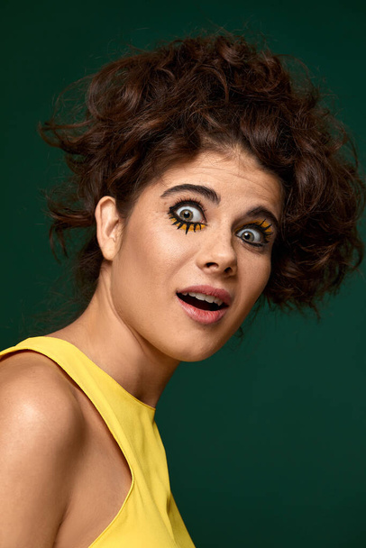 frightened curly woman with creative make-up in yellow dress on green background. - Photo, Image