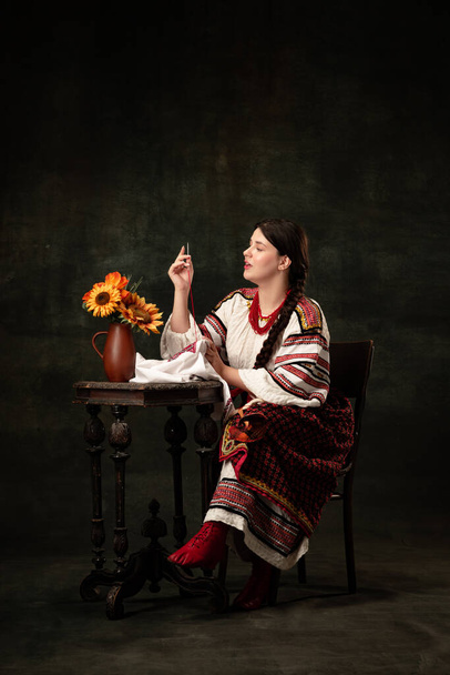 Cross stitching. Emotional adorable woman wearing national folk Ukrainian attire posing isolated over dark vintage background. Fashion, beauty, cultural heritage concept. - Photo, image