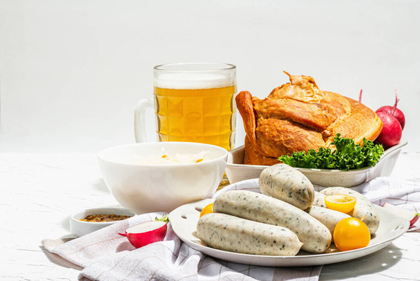Traditional Oktoberfest set. Beer, weisswurst, smoked pork knuckle eisbein with fermented cabbage. Festival food concept. Modern hard light, dark shadow, white plaster background, copy space - Фото, изображение