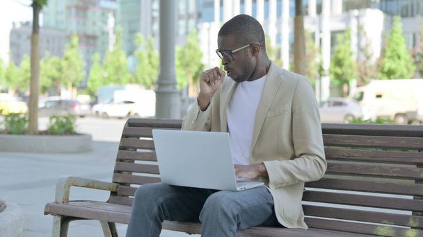 Tosse uomo afroamericano utilizzando il computer portatile mentre seduto all'aperto su panchina - Foto, immagini