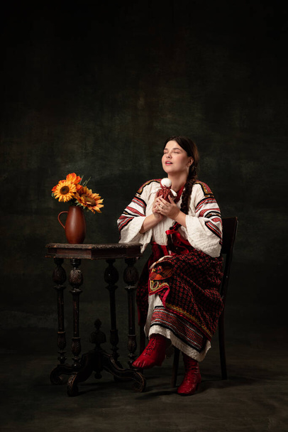 Fortune-telling. Young beautiful Ukrainian woman wearing traditional folk costume isolated over dark vintage background. Fashion, beauty, cultural heritage concept. Human emotions - 写真・画像