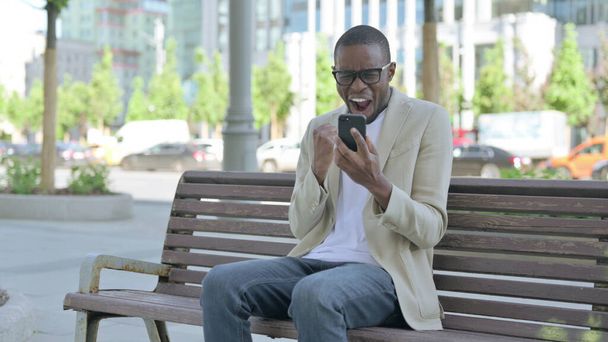 Afrikkalainen amerikkalainen mies juhlii online menestystä älypuhelimella istuessaan ulkona penkillä - Valokuva, kuva