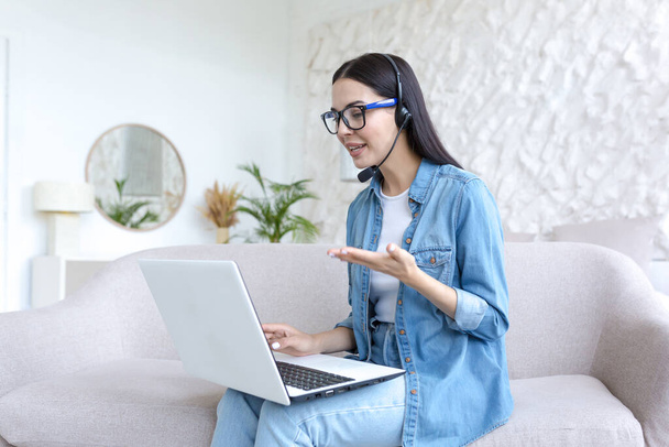 Online work. Young beautiful freelancer woman in glasses and jeans sitting at home on sofa and working with laptop and headphones online, talking on video call with clients, partners. - Photo, image