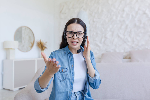 Webinar. Young beautiful business woman in glasses and headphones sits on the sofa at home and conducts a webinar, online training on camera, consults, tells, gestures with hands, smiles. - 写真・画像