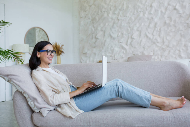 Online work. Young business woman in glasses, freelancer working at home on laptop. Lying comfortably on the couch in jeans and a shirt, typing - Fotografie, Obrázek