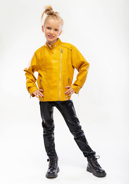 Pretty blonde little girl in a leather mustard jacket, black snake texture leggings and boots. High fashion, full length standing pose, isolated against a studio background - Photo, Image