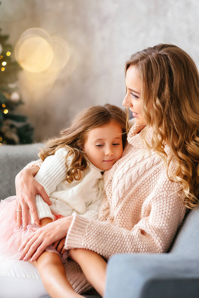 Mom and daughter on New Years Eve are sitting in a Scandinavian-style room in light warm sweaters with long flowing hair on a gray sofa, happy and hugging. High quality photo - Photo, image