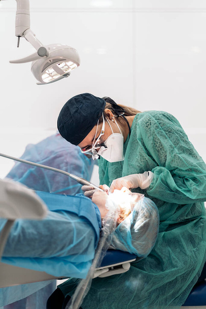 Female dentist wearing face mask and dental magnifying glasses doing treatment to unrecognized patient. - Foto, Imagen