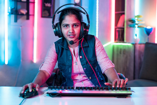 Smiling woman playing video game by talking on headphones while looking at camera at neon background - concept of tournament, entertainment and gaming addiction - Foto, imagen