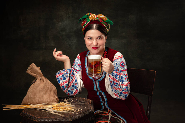 One young beautiful Ukrainian woman in traditional folk Ukrainian costume drinking beer isolated over dark vintage background. Fashion, beauty, cultural heritage, eras comparison concept. - Фото, зображення